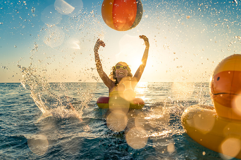 Ein jubelndes Kind mit gelber Sonnenbrille und Schwimmreifen wirft einen Wasserball in die Luft, während es im funkelnden Gegenlicht eines goldenen Sonnenuntergangs von Wassertropfen umspritzt wird. Packliste Sommerurlaub Wasser