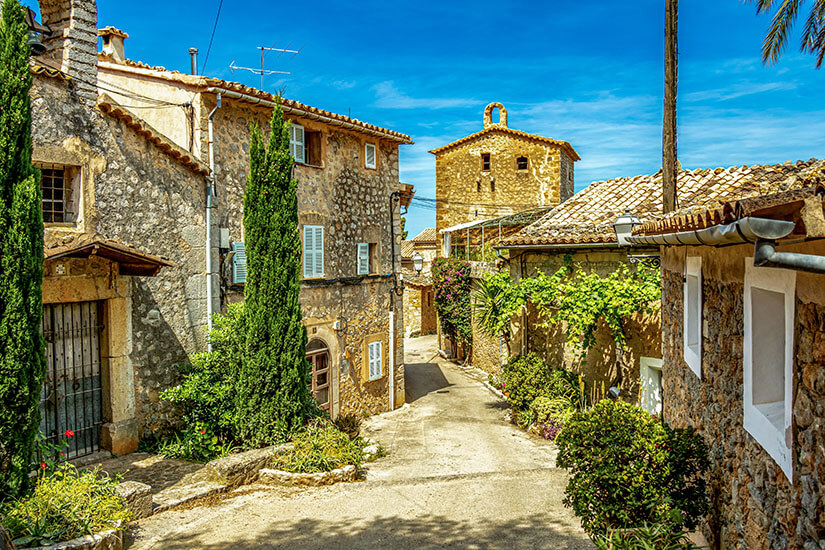 Gebäude aus hellem Stein und mit grüner Bepflanzung säumen einen hellen Weg, der durch das Dorf führt. Deia Spanien Willkommen