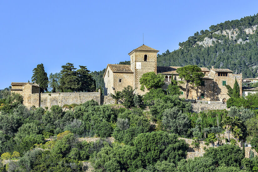 Eine Kirche aus hellem Stein thront auf einem Hügel, umgeben von grüner Vegetation. Deia Spanien Kirche