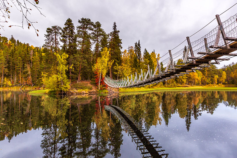 Eine hölzerne Hängebrücke spannt sich über einen ruhigen Fluss im Oulanka-Nationalpark, in dessen dunkler Oberfläche sich die Brücke und die herbstlich gefärbten Bäume am Ufer unter einem bewölkten Himmel spiegeln. Nationalparks Finnland Oulanka