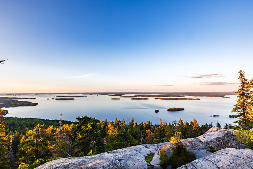 Blick von den hellen Felsen des Koli-Berges auf den weiten Pielinen-See mit seinen zahlreichen bewaldeten Inseln unter einem klaren, hellblauen Abendhimmel. Nationalparks Finnland Koli