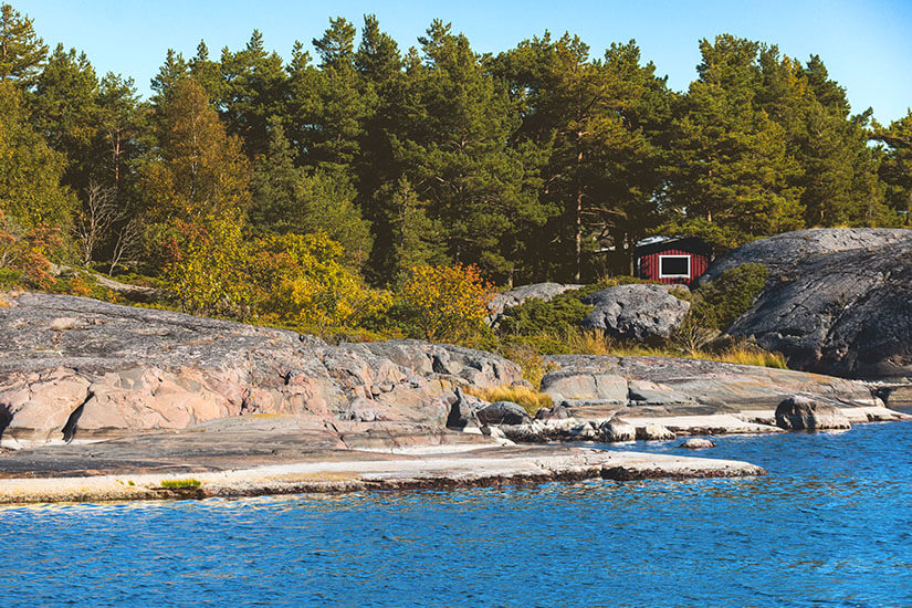 Glatte graue Felsen ragen am Ufer aus dem blauen Meer, dahinter steht eine kleine rote Hütte am Rand eines dichten Nadelwaldes unter einem klaren Himmel. Finnland Schaerenmeer Nationalparks Finnland Schaerenmeer
