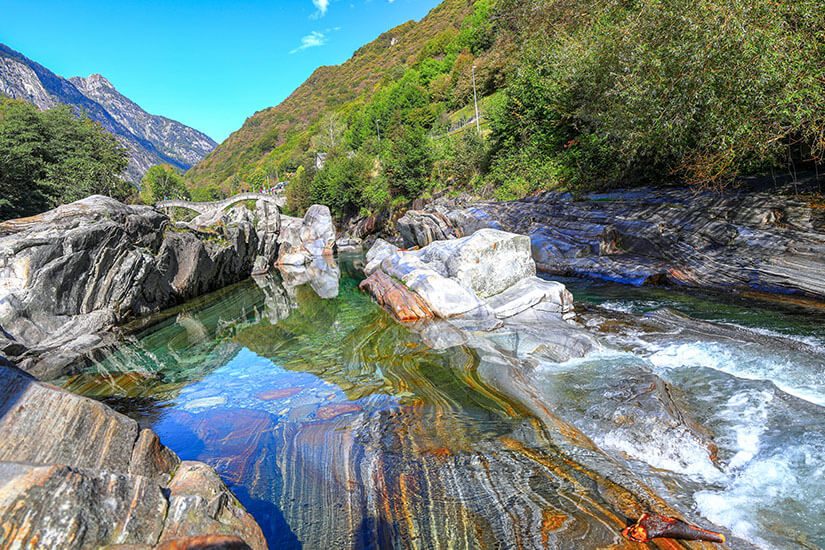 Die Verzasca fließt bei Lavertezzo über kunstvoll geschliffene Felsformationen, die die besondere Geologie des Tals sichtbar machen. Das kristallklare, smaragdgrüne Wasser lädt im Sommer zum Baden ein und zählt zu den beliebtesten Badestellen der Schweiz. Die vom Wasser polierten Gneisfelsen erzählen Jahrmillionen geologischer Geschichte und machen diesen Ort zu einem Highlight für Wandern und Baden im Verzascatal. Verzascatal Schweiz Fluss in Lavertezzo
