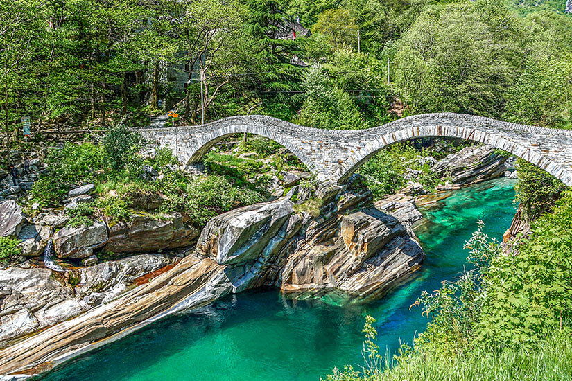 Die Ponte dei Salti ist die berühmteste Römerbrücke des Verzascatals und spannt sich mit ihrem doppelten Steinbogen über das smaragdgrüne Wasser der Verzasca. Die mittelalterliche Brücke bei Lavertezzo ist das Wahrzeichen der Valle Verzasca und eine der meistbesuchten Sehenswürdigkeiten im Tessin. Unterhalb der Brücke befinden sich die beliebtesten Badestellen des Tals, wo Mutige von den Felsen ins kristallklare Wasser springen. Verzascatal Schweiz Ponte dei Salti
