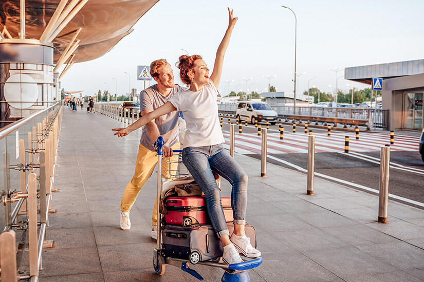 Ein junges Pärchen tobt ausgelassen vor einem Flughafenterminal: Die Frau sitzt jubelnd auf einem mit Koffern beladenen Gepäckwagen, während ihr Begleiter ihn lachend schiebt und dabei eine freche Pose hinlegt. Cashback Reisebuchung Paar Flughafen