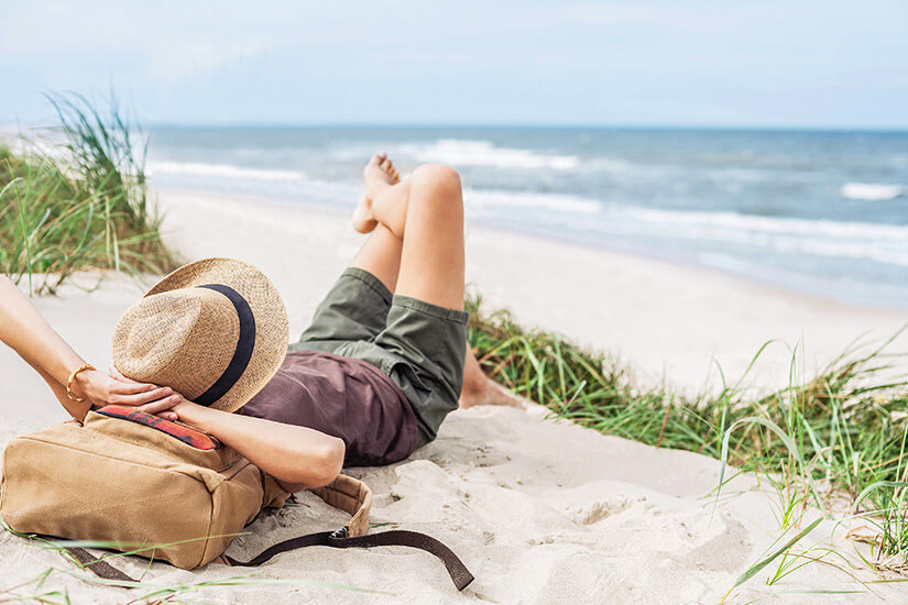 Von Strandhafer umgeben liegt eine Urlauberin entspannt in den Dünen mit einem Strohhut über dem Gesicht und einem Rucksack als Kopfkissen. Cashback Reisebuchung Strand