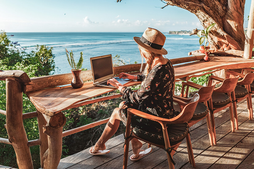 Eine junge Frau mit Strohhut arbeitet an ihrem Laptop an einem rustikalen Holztisch auf einer offenen Terrasse, während sie einen atemberaubenden Blick über das türkisblaue Meer genießt. Cashback Reisebuchung Laptop