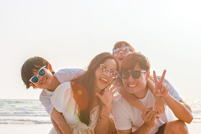 Eine fröhliche vierköpfige Familie mit stylischen Sonnenbrillen und weißen T-Shirts posiert ausgelassen am Strand, wobei die Kinder auf dem Rücken der lachenden Eltern sitzen und alle gemeinsam in die Kamera strahlen. Cashback Reisebuchung Familie