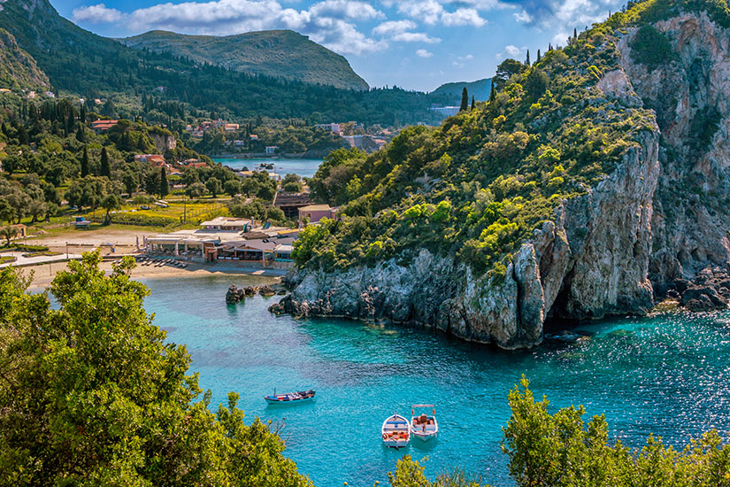 Eine malerische Bucht mit kristallklarem türkisfarbenen Wasser, von bewaldeten Felsen und üppiger mediterraner Vegetation eingerahmt, beherbergt zwei kleine Boote, während im Hintergrund ein sandiger Strand mit Liegestühlen und eine von Bergen gekrönte Ortschaft zu erkennen sind. Kos oder Korfu Griechenland