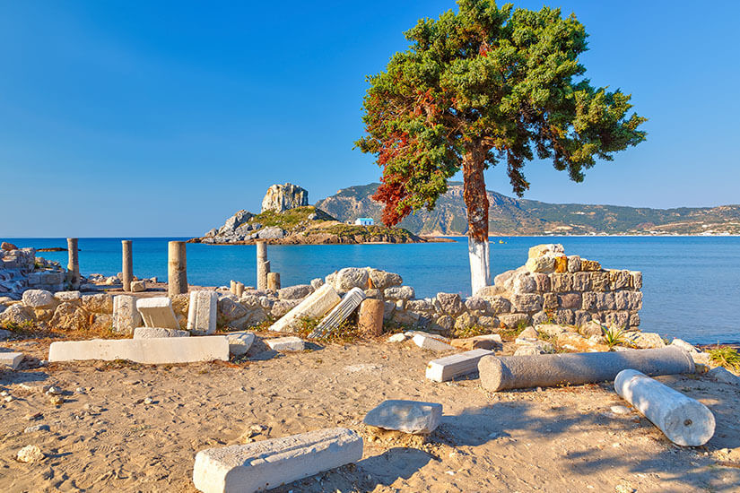 Verstreute Marmorsäulen und antike Mauerreste einer frühchristlichen Basilika liegen im sandigen Küstengelände unter einem markanten Baum, während im Hintergrund eine kleine Felsinsel mit einer blauen Kapelle im tiefblauen Ägäischen Meer auftaucht. Kos oder Korfu Ruinen