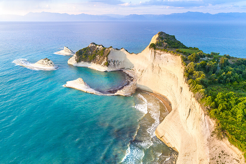 Eine spektakuläre Luftaufnahme zeigt dramatische, cremeweiße Kalksteinklippen, die steil ins türkisblaue Ionische Meer abfallen und eine abgeschiedene kleine Bucht mit vorgelagerten Felsformationen einrahmen. Kos oder Korfu Drastis