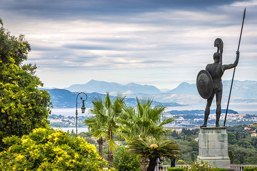 Die imposante Bronzestatue des Achilles mit Speer und Schild überragt die üppigen Gärten des Achilleion-Palastes auf Korfu und bietet einen weiten Panoramablick über die Küstenstadt, das Meer und die albanischen Berge im Hintergrund. Kos oder Korfu Achilleion