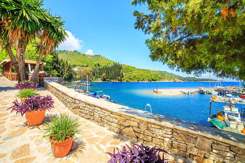 Eine gepflasterte Uferpromenade mit bunten Topfpflanzen und schattenspendenden Bäumen säumt einen malerischen kleinen Fischerhafen mit tiefblauem Wasser, vertäuten Booten und bewaldeten Hügeln im Hintergrund. Kos oder Korfu Kouloura