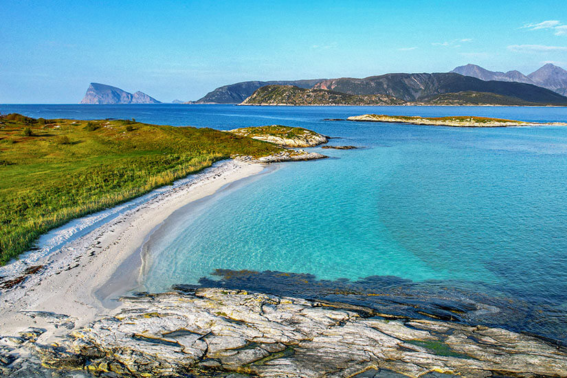 Ein karibisch anmutender Senja Strand mit weißem Sand und unglaublich türkisem Wasser – und das weit nördlich des Polarkreises. Grüne Wiesen reichen bis ans Ufer, im Hintergrund zeichnen sich sanfte Bergsilhouetten ab. Dieser Strand beweist, dass die Senja Strände zu den schönsten und überraschendsten Küstenabschnitten ganz Norwegens gehören. Senja Norwegen Strand