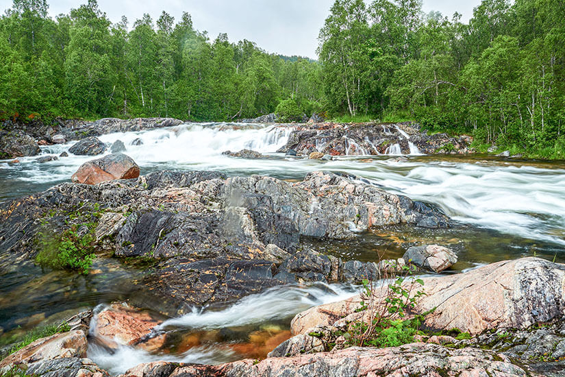 Der Anderdalen Nationalpark auf Senja zeigt sich hier von seiner wildesten Seite: Ein reißender Fluss bahnt sich seinen Weg über glattgeschliffene Felsen, umgeben von sattem Birkenwald. Die Langzeitbelichtung verleiht dem Wasser eine seidig-weiche Textur, die den Kontrast zu den rauen Steinen wunderbar betont. Dieses Naturjuwel gehört zu den schönsten Senja Sehenswürdigkeiten und ist ein Paradies für alle, die unberührte Wildnis suchen. Senja Norwegen Anderdalen National Park