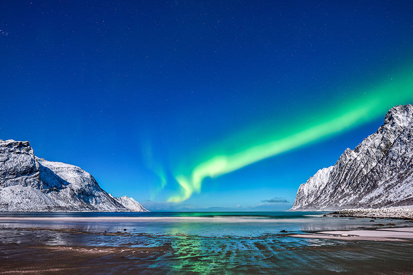 Grün leuchtende Nordlichter tanzen über den Nachthimmel von Senja und spiegeln sich im eiskalten Wasser eines winterlichen Fjords. Schneebedeckte Berge rahmen die Szenerie ein und sorgen für eine fast überirdische Atmosphäre. Senja in winter ist einer der besten Orte Norwegens, um die Aurora Borealis zu erleben. Senja Norwegen Polarlichter