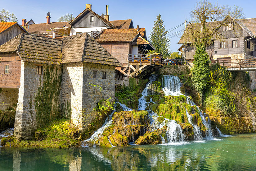 Ein Wasserfäll läuft inmitten eines Dorfes über grüne Felsen in ein Wasserbecken. Wasserfaelle Kroatien Willkommen