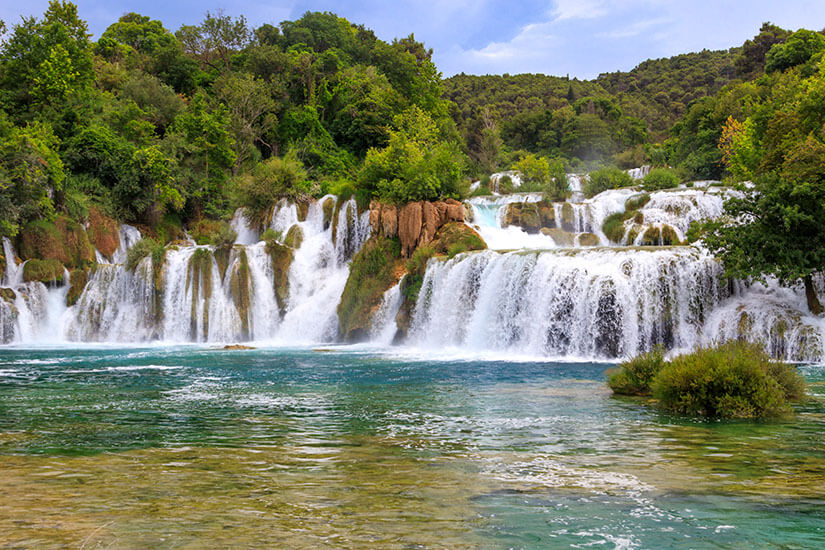 Weiße breite Wasserfälle ergießen sich über rot-braunes Gestein in einen flachen See, der in Türkistönen schimmert. Wasserfaelle Kroatien Skradinski