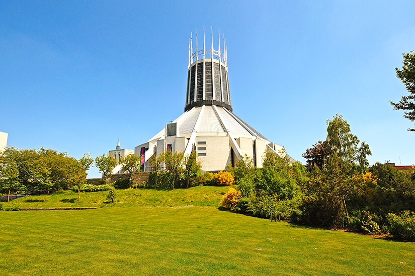 Die moderne, zeltartige Liverpool Metropolitan Cathedral mit ihrem markanten Laternenturm aus Glas und Metall erhebt sich hinter einer grünen Wiese und Bäumen unter einem strahlend blauen Himmel. Liverpool England Metropolitan Cathedral