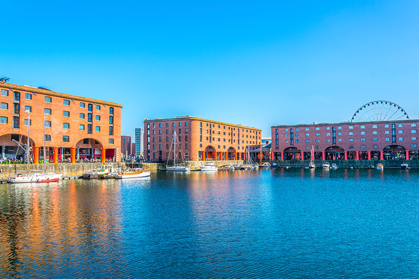 Die historischen Backsteingebäude des Royal Albert Dock in Liverpool mit ihren markanten roten Säulen säumen das Hafenbecken, in dem mehrere Segelboote vor einem Riesenrad im Hintergrund ankern. Liverpool England Royal Albert Dock