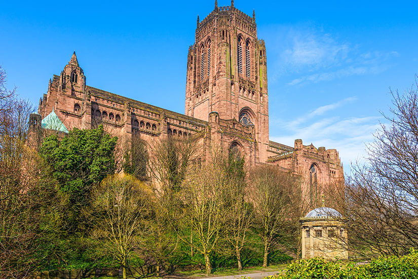 Die monumentale Liverpool Cathedral aus rotem Sandstein mit ihrem imposanten zentralen Turm erhebt sich hinter einer Anhöhe mit teils belaubten, teils kargen Bäumen und einem kleinen Pavillon unter blauem Himmel. Liverpool England Cathedral