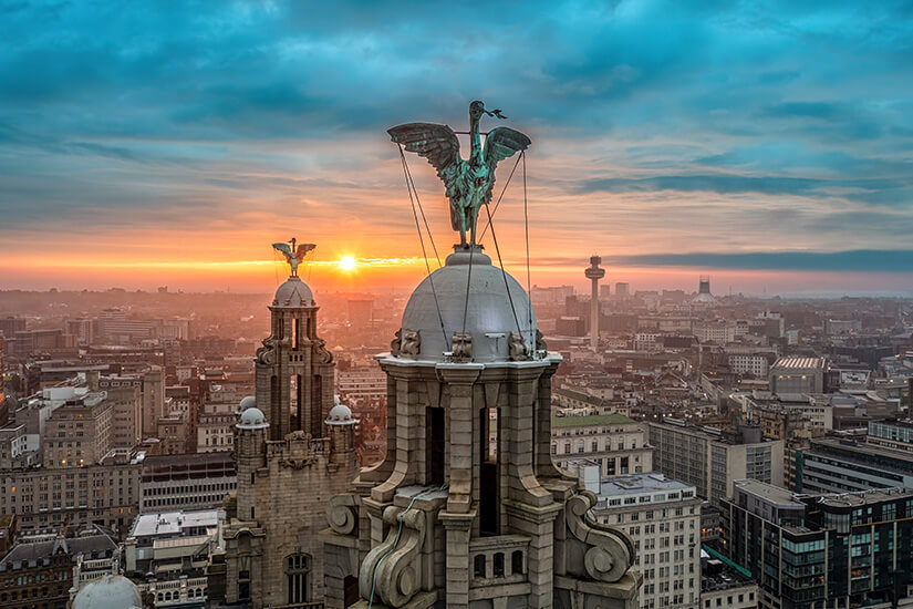 Nahaufnahme der Turmspitze des Royal Liver Building in Liverpool mit einem kupfernen Liver Bird vor einer dämmrigen Stadtlandschaft und einem farbenfrohen Sonnenuntergang am Horizont. Liverpool England Royal Liver Building