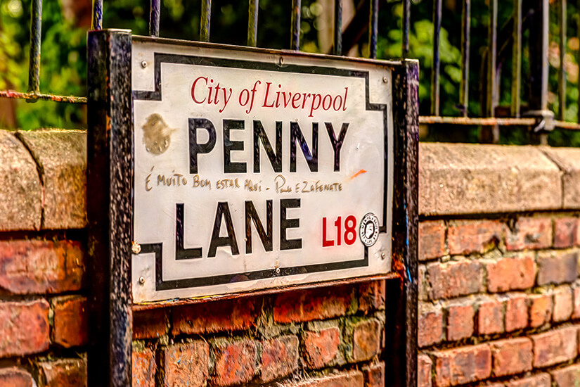Ein weißes Straßenschild mit der schwarzen Aufschrift „PENNY LANE“ und dem roten Postleitzahlbezirk „L18“ ist an einer alten Backsteinmauer befestigt, wobei handschriftliche Notizen von Fans auf dem Schild zu sehen sind. Liverpool England Penny Lane
