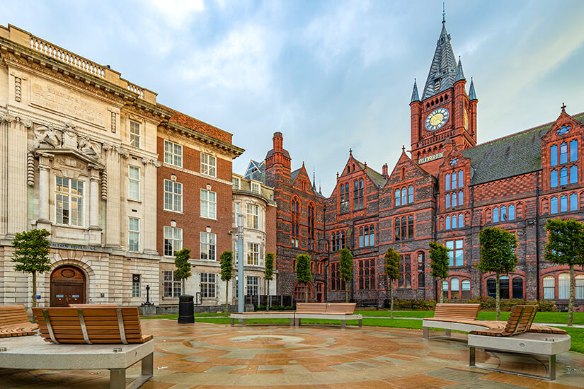 Das beeindruckende neugotische Gebäude der Victoria Gallery aus rotem Backstein mit seinem hohen Uhrenturm steht an einem gepflasterten Platz mit modernen Holzbänken und kleinen Bäumen unter einem bewölkten Himmel. Liverpool England Victoria Gallery