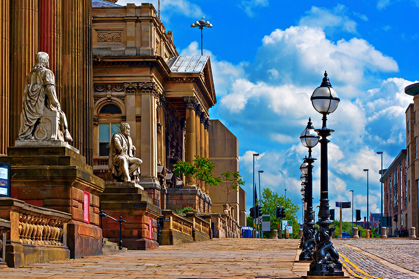 Eine gepflasterte Straße in Liverpool, gesäumt von prachtvollen neoklassizistischen Gebäuden mit hohen Säulen und Steinstatuen auf der linken Seite sowie einer Reihe historischer schwarzer Laternenpfähle unter einem blauen Himmel mit weißen Wolken. Liverpool England William Brown Street