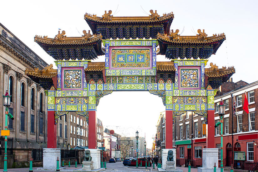 Das prachtvolle, farbenfroh verzierte Eingangstor zur Chinatown in Liverpool mit roten Säulen, goldenen Drachenfiguren und traditionellen blauen Dächern überspannt eine Straße unter hellem Himmel. Liverpool England Chinatown