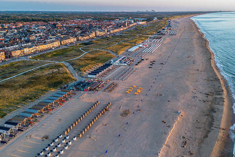 Die beeindruckende Luftaufnahme zeigt den weitläufigen Strand von Katwijk aan Zee mit seinen typischen bunten Strandhäuschen und der dahinterliegenden Promenade. Kilometerweit erstreckt sich der feine Sandstrand entlang der Küste und bietet Platz für zahllose Erholungssuchende. Die Stadt mit ihren charakteristischen roten Dächern schmiegt sich harmonisch zwischen Dünen und Meer und zählt zu den beliebtesten Küstenorten der Niederlande. Katwijk aan Zee Niederlande Strand