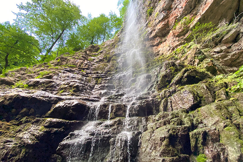Ein Wasserfall fällt von einer braunen Felswand herunter. Grüne Bäume umgeben die Szene. Wasserfaelle Kroatien Zeleni