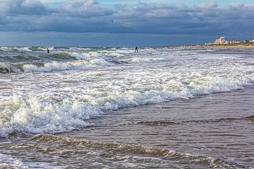 Die raue Nordsee vor Katwijk aan Zee zeigt sich hier von ihrer kraftvollen Seite mit hohen Wellen, die das Herz jedes Wassersportlers höherschlagen lassen. Surfer und Kitesurfer nutzen die windigen Tage, um auf dem Meer ihrem Sport nachzugehen, während sich am Horizont die Silhouette der Stadt abzeichnet. Der Strand von Katwijk ist gerade bei frischem Wetter ein beliebter Spot für Aktivurlauber in den Niederlanden. Katwijk aan Zee Niederlande Meer