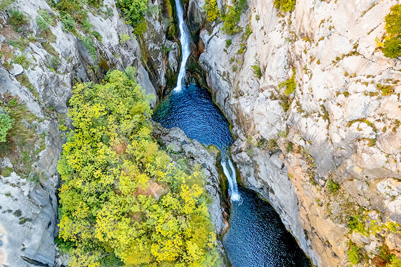 Ein Wasserfall in einer Schlucht aus der Vogelperspektive gesehen. Weiße Kaskaden rauschen in blaues Wasser. Grüne Vegetation umgibt die Felsen. Wasserfaelle Kroatien Gubavica