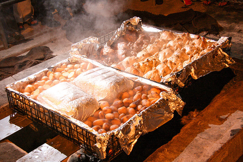 Große, mit Alufolie ausgekleidete Metallroste sind mit ganzen Kartoffeln und Hühnchenstücken beladen, die unter aufsteigendem Dampf garen und auf eine traditionelle Erdofen-Garmethode hindeuten. Neuseeland Essen Hangi