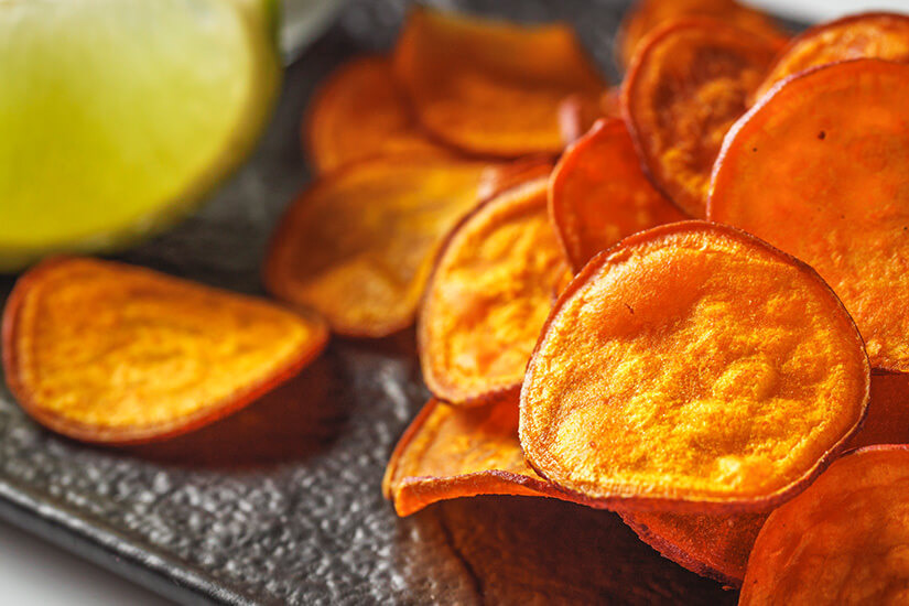 Knusprig gebackene Süßkartoffelchips in leuchtendem Orange-Rot sind auf einem dunklen Serviertablett angerichtet, während im Hintergrund eine angeschnittene grüne Limette als frische Geschmackskomponente zu erkennen ist. Neuseeland Essen Kumara Chips
