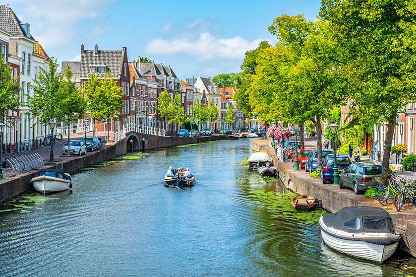 Ein ruhiger Kanal zieht sich durch eine Stadt mit historischen Häusern entlang des Ufers. Kleine Boote liegen am Rand oder fahren über das Wasser, während Bäume die Straßen säumen. Fahrräder und parkende Autos stehen entlang der Promenade. Leiden Stadt