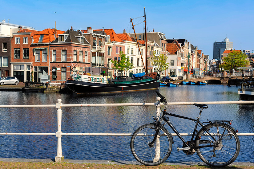 Ein Fahrrad steht an einem Geländer direkt an einem Kanalufer. Auf dem Wasser liegt ein größeres Boot, dahinter reihen sich typische Stadthäuser mit Ziegeldächern. Eine kleine Brücke überspannt den Kanal. Holland Rad