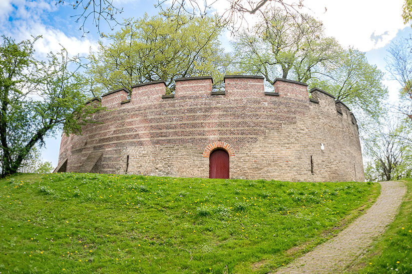 Eine runde, festungsartige Backsteinmauer erhebt sich auf einem kleinen Hügel. Ein schmaler Weg führt hinauf zu einer roten Tür in der Mauer. Oben wachsen Bäume, die über den Rand hinausragen und die Anlage umgeben. Leiden Burcht