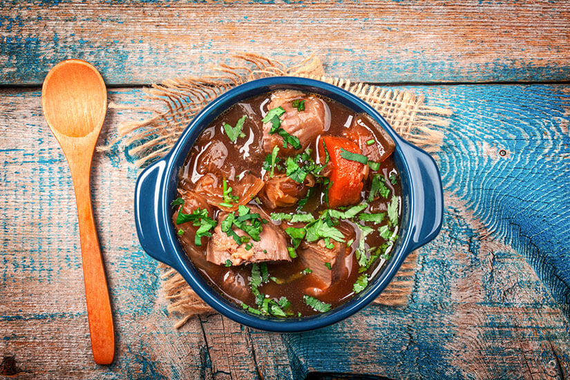 Das rustikale Holzbrett und der einfache Holzlöffel unterstreichen den bodenständigen Charakter dieses Schmorgericht-Klassikers. Neuseeland Essen Lamm Eintopf