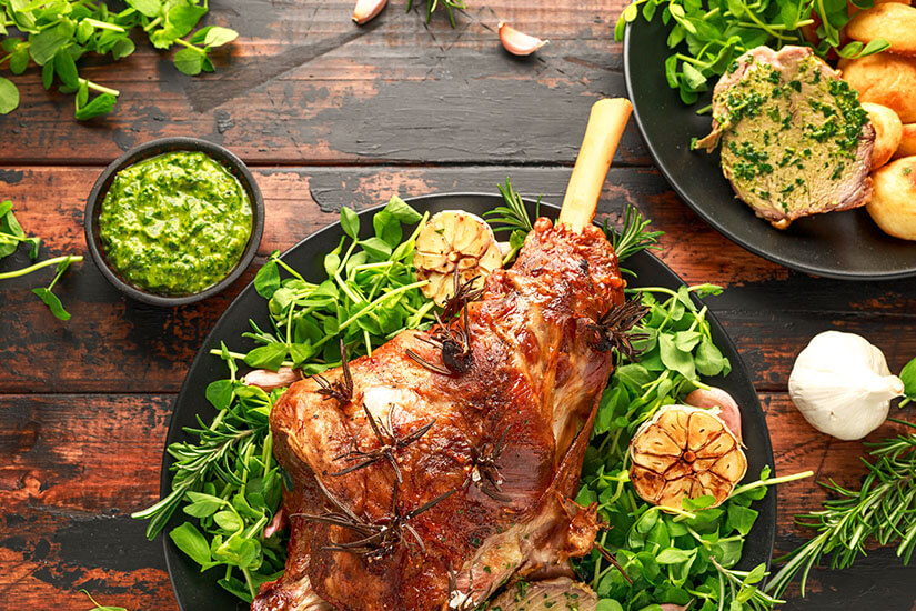 Eine prächtig gebratene Lammkeule mit Knochen, gespickt mit Rosmarinzweigen und umgeben von geröstetem Knoblauch und frischem Feldsalat, thront als Mittelpunkt einer festlichen Tafel, flankiert von einer leuchtend grünen Soße und goldbraunen Bratkartoffeln. Neuseeland Essen Lamm