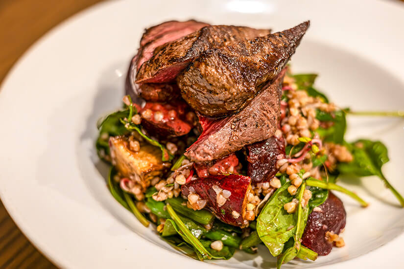 Zart rosa gebratene Fleischscheiben thronen auf einem ansprechend angerichteten Bett aus Rucola, gekochtem Buchweizen und gerösteten Roten Beten, das einem gehobenen Restaurantgericht würdig ist. Neuseeland Essen Hirsch