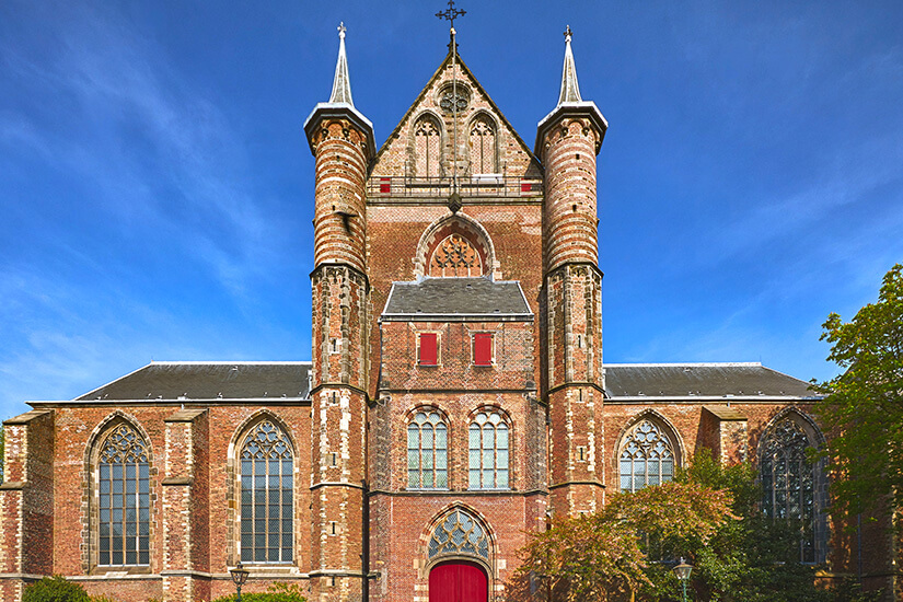 Eine große Backsteinkirche mit gotischen Elementen und hohen, schmalen Fenstern dominiert die Szene. Zwei Türme rahmen die Fassade ein, darüber ragt ein spitzes Dach mit Kreuz empor. Vor dem Gebäude stehen Bäume. Leiden Kirche