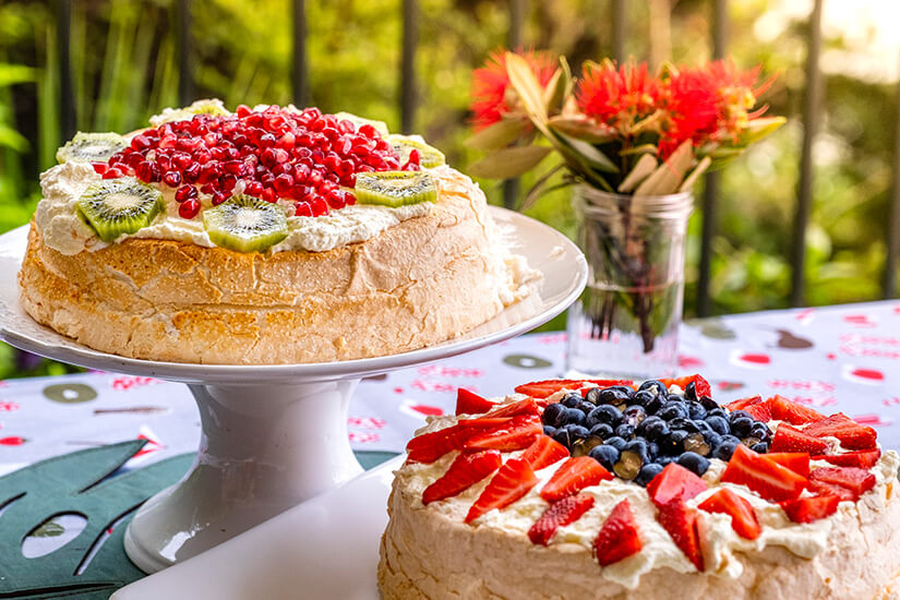 Zwei luftige Pavlova-Torten mit knuspriger Baiserhülle und Schlagsahne sind festlich mit frischen Früchten dekoriert und stehen auf einem sommerlich gedeckten Tisch im Freien. Neuseeland Essen Pavlova