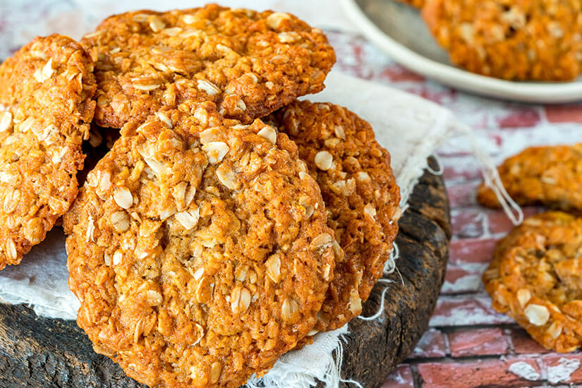Goldbraun gebackene Haferflocken-Cookies mit ihrer charakteristisch rauen, rustikalen Oberfläche sind auf einem Leinentuch auf einem Holzbrett gestapelt und verströmen das Flair selbstgebackener Hausmannskost. Neuseeland Essen ANZAC