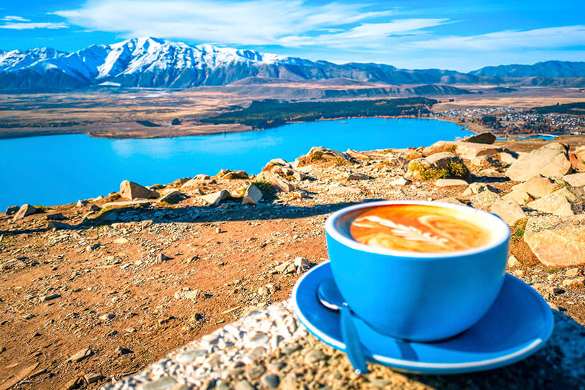 Ein kunstvoll zubereiteter Kaffee mit Milchschaummuster in einer blauen Tasse thront im Vordergrund, während sich dahinter das atemberaubende Panorama des leuchtend türkisblauen Lake Tekapo mit seinen schneebedeckten Südalpengipfeln auf der Südinsel Neuseelands entfaltet. Neuseeland Essen Kaffee