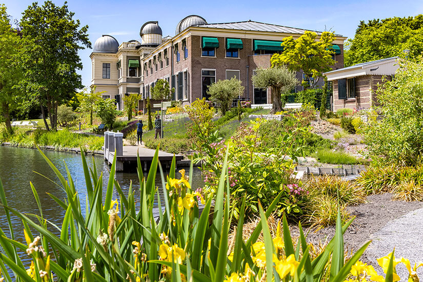Ein gepflegter Garten mit blühenden Pflanzen liegt an einem kleinen Gewässer. Dahinter steht ein größeres Gebäude mit Kuppeln, das an ein Observatorium oder wissenschaftliches Institut erinnert. Wege und Terrassen führen durch die Anlage, auf denen sich einzelne Personen bewegen. Leiden Botanischer Garten
