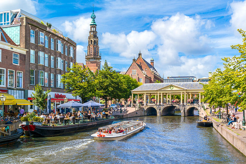 Eine belebte Grachtenlandschaft mit Booten auf dem Wasser und vielen Menschen an den Ufern. Entlang des Kanals stehen historische Gebäude, Cafés und Terrassen mit Gästen. Eine Brücke mit Bögen überspannt den Kanal, dahinter ragt ein Turm in den Himmel. Leiden Grachten