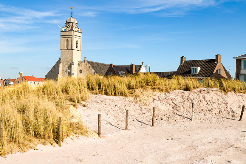 Sandige Dünen mit Strandgras ziehen sich durch den Vordergrund, durchzogen von einem einfachen Zaun. Dahinter stehen einige Häuser und ein markanter Kirchturm mit Uhr. Leiden Strand