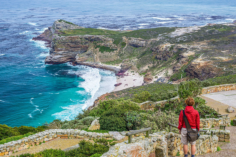 Eine Person mit roter Jacke blickt von einem Pfad gesäumt von einer niedrigen Steinmauer auf eine Atlantikküste herunter. Weiße Wellen erreichen den Strand. Suedafrika Nationalparks Aussicht